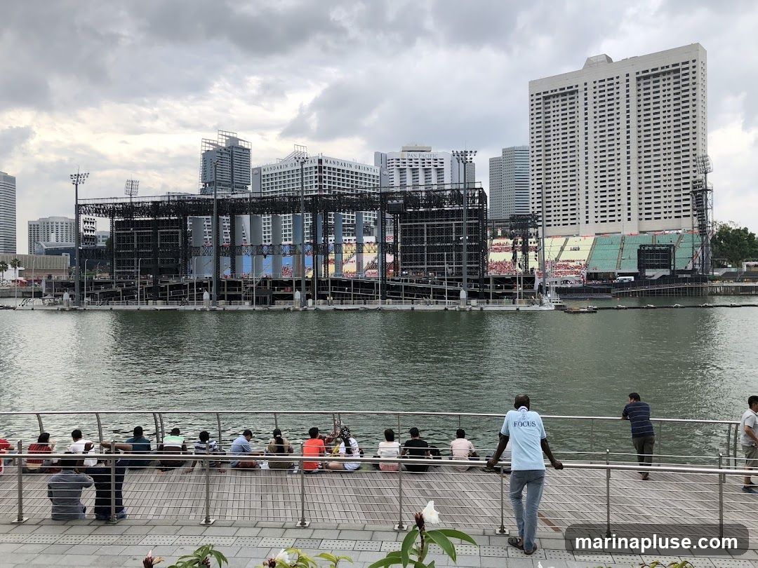 A group of people are seated on a wooden boardwalk, facing a large outdoor stage structure across a body of water. The stage appears to be under construction or set up for an event, with scaffolding and screens visible. In the background, several tall buildings form a city skyline under a cloudy sky.