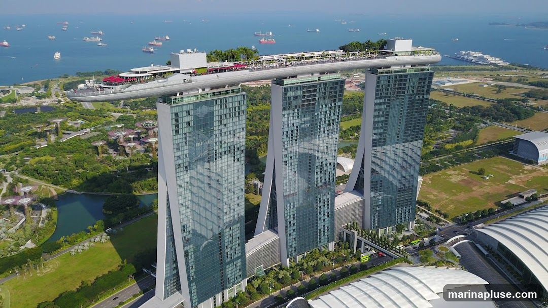 An aerial view of the Marina Bay Sands hotel in Singapore, with its iconic three towers topped by a ship-like structure. The hotel is situated next to Gardens by the Bay, a nature park featuring futuristic Supertree structures. The Singapore Strait, dotted with numerous cargo ships, is visible in the background.