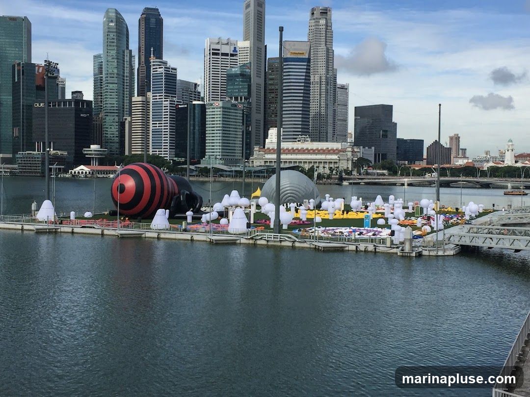 A wide shot of a body of water with a floating platform in the foreground. The platform is adorned with numerous inflatable sculptures, including a large red and black striped insect-like figure and many smaller white, bulbous shapes. In the background, a dense city skyline with numerous skyscrapers rises against a cloudy sky. The water is calm and reflects the sky and buildings.