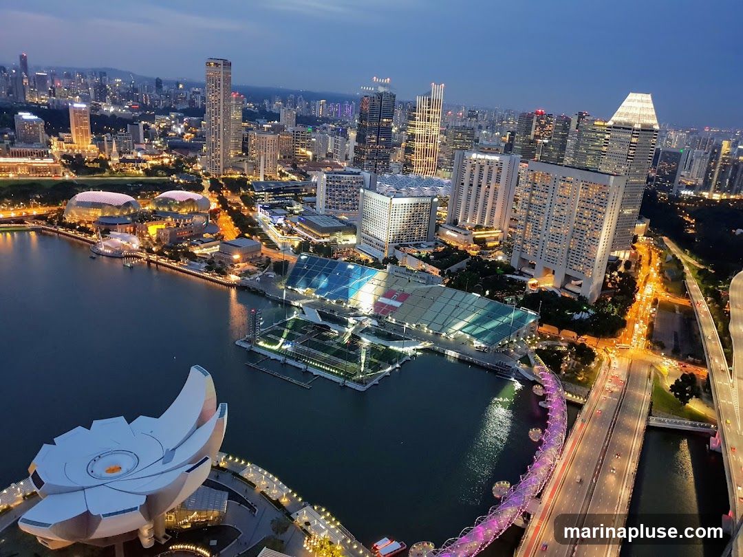 An aerial view of the Singapore skyline at dusk, showcasing the Marina Bay area. The image features the iconic Marina Bay Sands hotel, the ArtScience Museum with its lotus-like design, and the Esplanade – Theatres on the Bay. Illuminated bridges and highways are visible, along with numerous skyscrapers and city lights stretching into the distance. The water of Marina Bay reflects the city lights, creating a vibrant urban panorama.