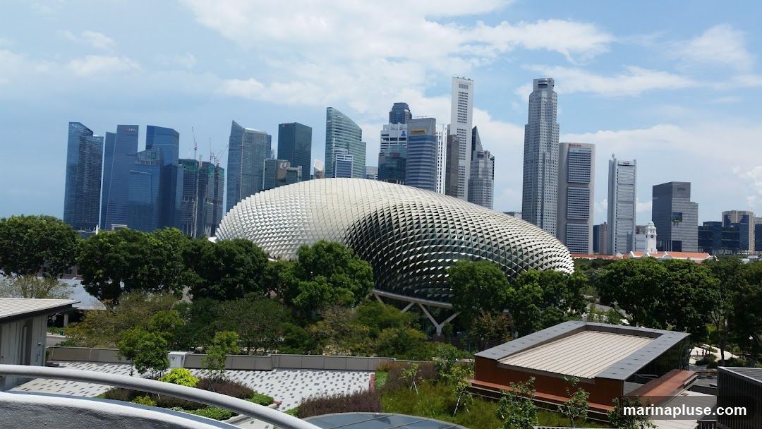 A panoramic view of the Singapore skyline, featuring numerous modern skyscrapers and the distinctive durian-shaped Esplanade - Theatres on the Bay. Lush green trees and landscaping are visible in the foreground, with parts of a modern building structure in the lower right corner. The sky is partly cloudy.