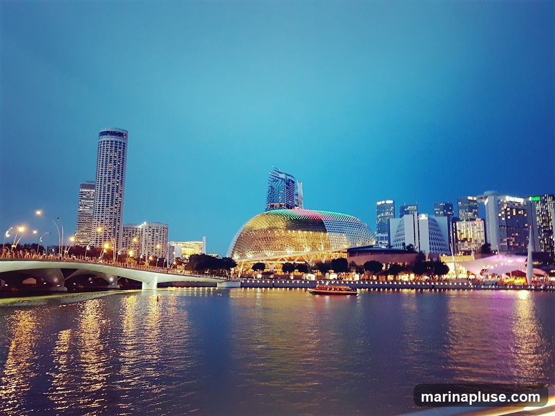 A wide shot of the Singapore skyline at dusk, with the iconic Marina Bay Sands and the Esplanade concert hall illuminated. A bridge spans the water in the foreground, with a boat sailing by. The city lights reflect on the water, creating a vibrant and colorful scene.