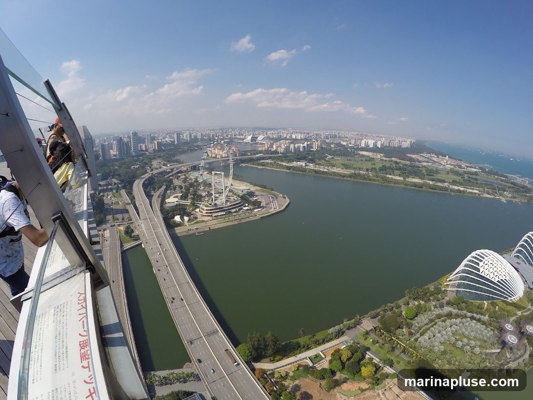 A wide-angle, high-angle view of the Singapore skyline on a clear day. In the foreground, a glass barrier with informational text in Japanese is visible, with two people standing near it. The view overlooks a large body of water, a highway with traffic, and the iconic Supertrees at Gardens by the Bay. In the distance, the city's skyscrapers and the Singapore Flyer Ferris wheel are prominent. The sea and a hazy horizon complete the scene.