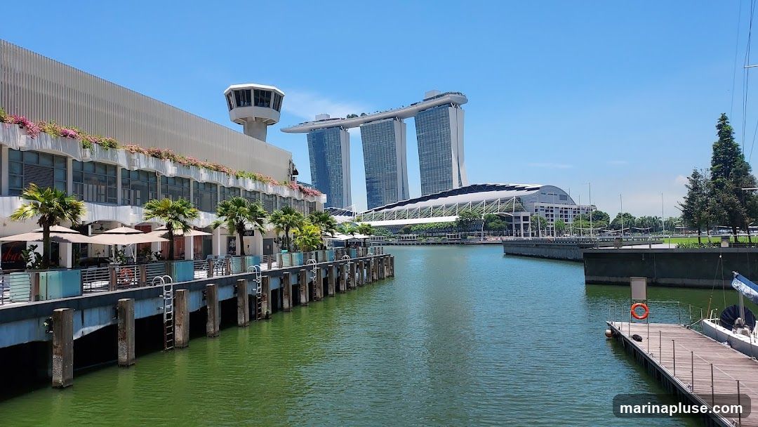 A scenic view of a waterfront promenade with outdoor seating areas and palm trees. In the background, the iconic Marina Bay Sands hotel and convention center are visible under a clear blue sky. A pier with boats is in the foreground.