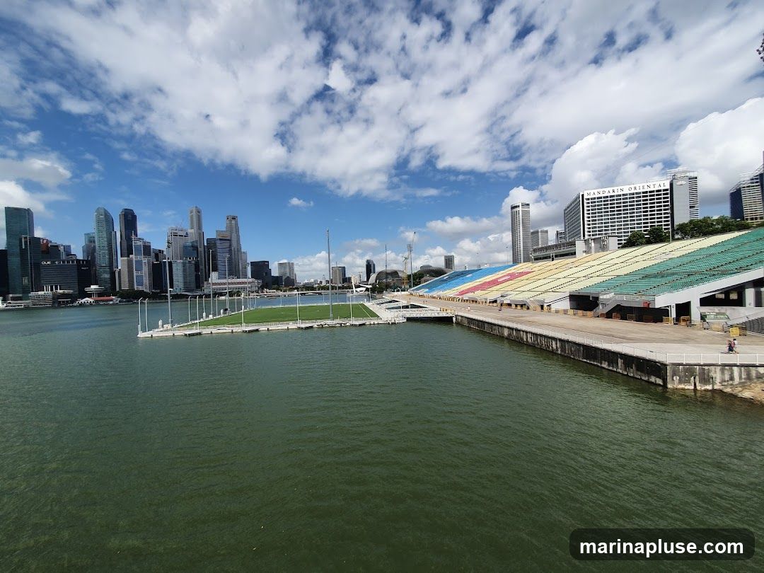 A wide shot of the Marina Bay area in Singapore, featuring the stadium with colorful seating in the foreground and the city skyline in the background. The water in the bay is a deep green, and the sky is blue with scattered white clouds. The stadium has sections of yellow, blue, and green seating. The Mandarin Oriental hotel is visible among the skyscrapers.