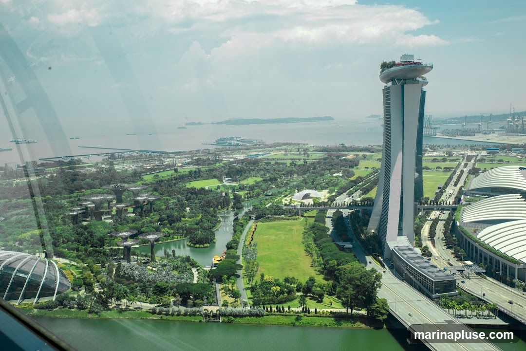 An aerial view of Singapore's iconic skyline, featuring the Marina Bay Sands hotel and the Supertree Grove at Gardens by the Bay. The image captures the urban landscape with lush greenery, waterways, and the vast expanse of the sea in the background, dotted with ships. The perspective is from a high vantage point, likely a building or observation deck, looking out over the city.