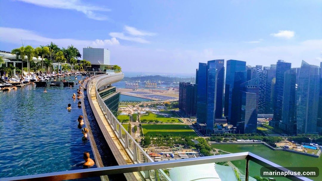 A panoramic view of a large infinity pool filled with people, set against a backdrop of a modern cityscape. Palm trees and lounge chairs line the edge of the pool. In the distance, skyscrapers dominate the skyline, with a harbor and port visible beyond. The sky is clear and blue with a few scattered clouds.