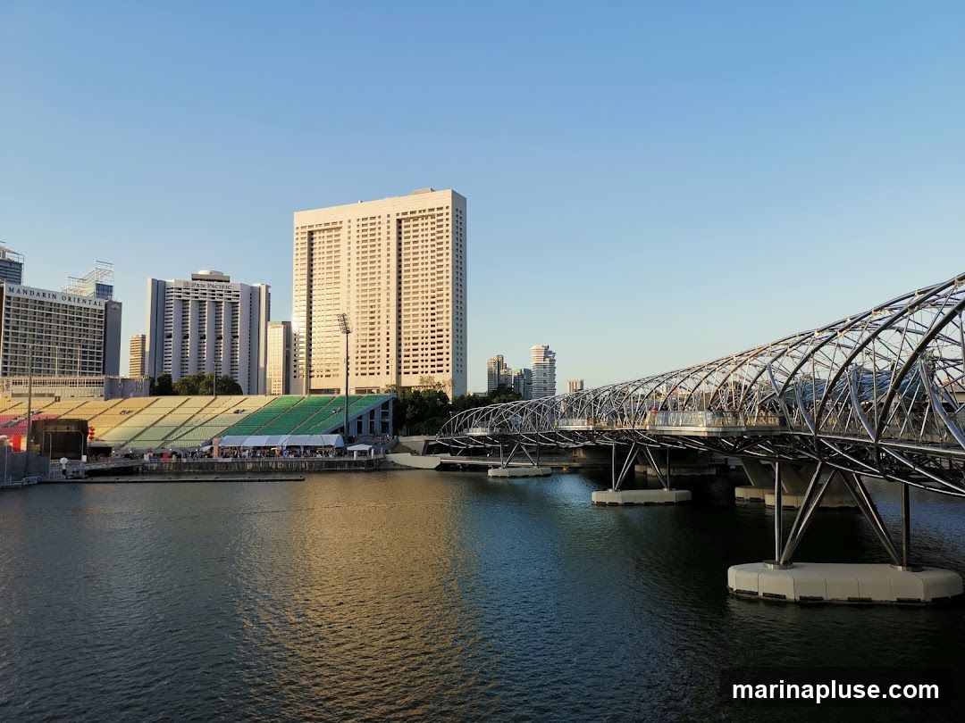 A wide shot of Marina Bay in Singapore, featuring the Helix Bridge in the foreground and several prominent buildings in the background, including the Mandarin Oriental and Pan Pacific hotels, as well as a stadium with tiered seating. The water reflects the clear blue sky and the golden hues of the setting sun.