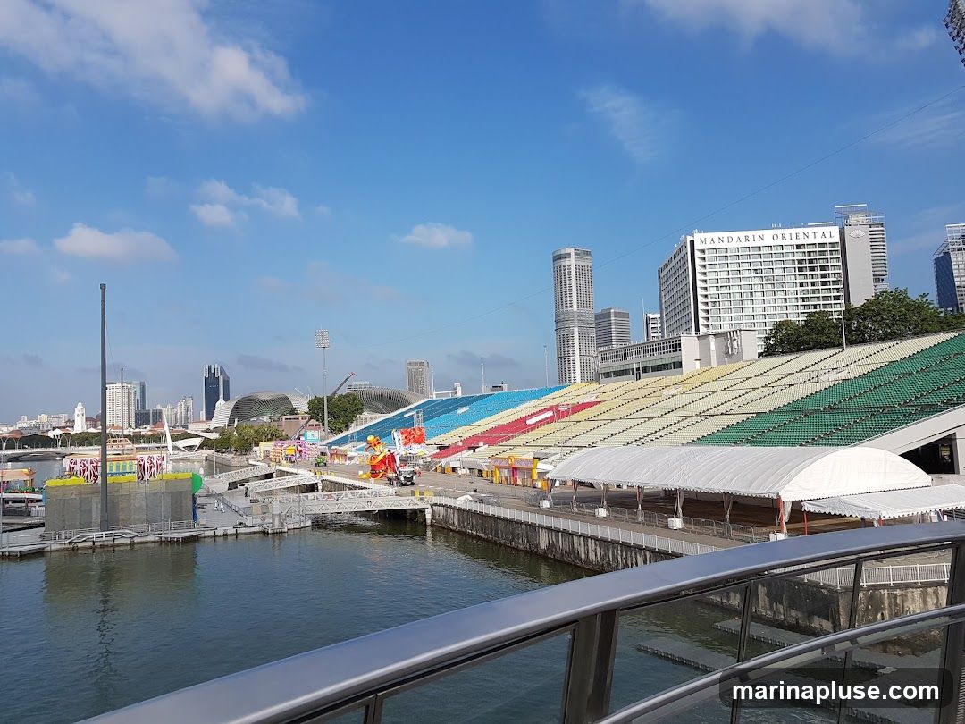 A wide shot of a waterfront area with stadium seating in the foreground and a city skyline in the background. The stadium seating is colorful, with sections in green, yellow, red, and blue. A white tent structure is set up along the waterfront. In the distance, various skyscrapers and buildings form the city skyline, including a prominent hotel labeled 'MANDARIN ORIENTAL'. The water in the foreground reflects the sky and buildings. There are some temporary structures and decorations visible along the waterfront, suggesting an event or festival.
