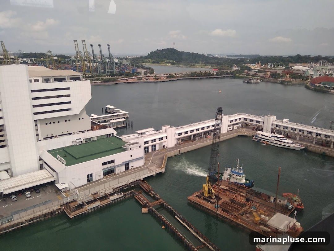 An aerial view of a busy harbor with large cranes in the background and various boats and barges in the foreground. A white building complex with a green rooftop is visible on the left, adjacent to the water. A long, white building with many windows lines the waterfront, with a large white yacht docked beside it. Construction barges and tugboats are also present in the water. The scene suggests a port or industrial area with ongoing maritime activity.