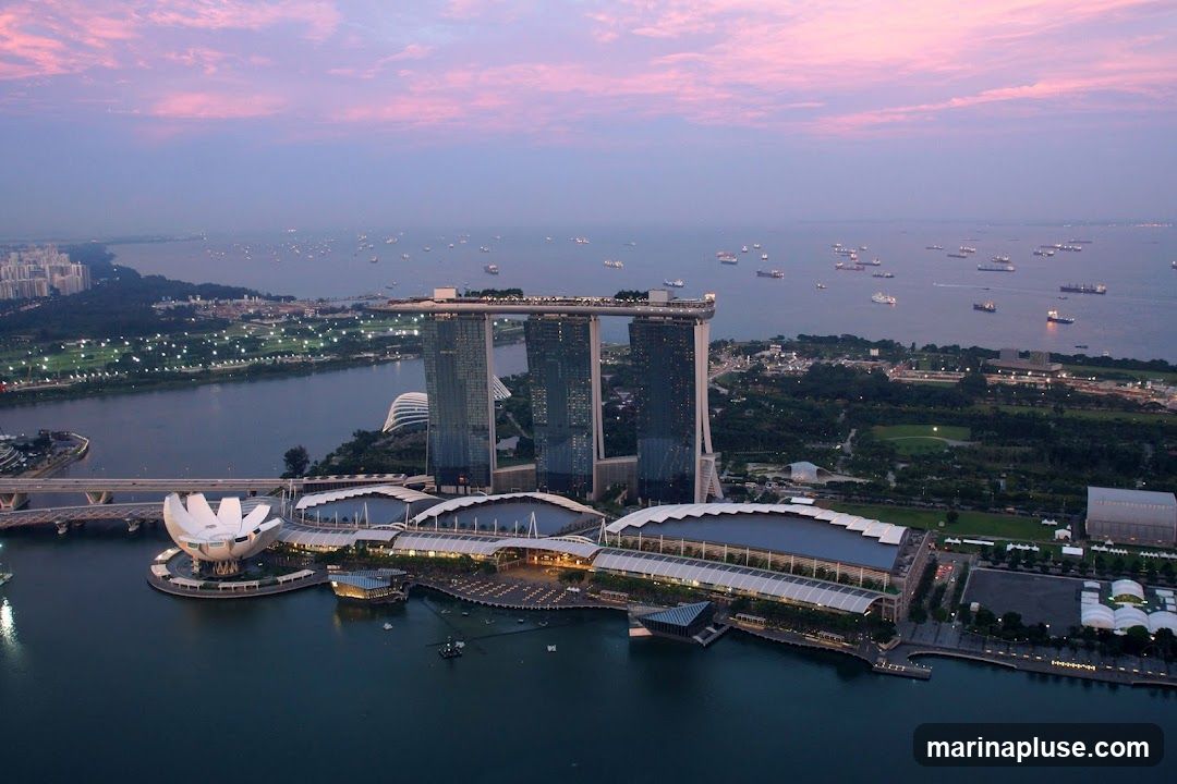 An aerial view of the Marina Bay Sands complex in Singapore during twilight. The iconic three towers, connected by a rooftop deck, are illuminated. In the foreground, the ArtScience Museum, shaped like a lotus flower, is visible. The surrounding waters are dotted with numerous ships. The sky displays soft hues of pink and purple from the setting sun.