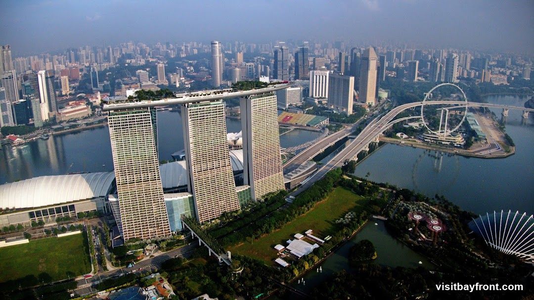 An aerial view of the Singapore skyline, featuring the iconic Marina Bay Sands hotel with its distinctive three towers and connecting skybridge. In the foreground, the Gardens by the Bay with its Supertree Grove and conservatories are visible. The Singapore Flyer Ferris wheel can be seen in the distance, along with a sprawling cityscape of numerous skyscrapers and a body of water.