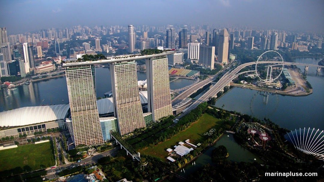 An aerial view of the Singapore skyline, featuring the iconic Marina Bay Sands hotel with its distinctive three towers and connecting skybridge. In the foreground, the Gardens by the Bay with its Supertree Grove and conservatories are visible. The Singapore Flyer Ferris wheel can be seen in the distance, along with a sprawling cityscape of numerous skyscrapers and a body of water.