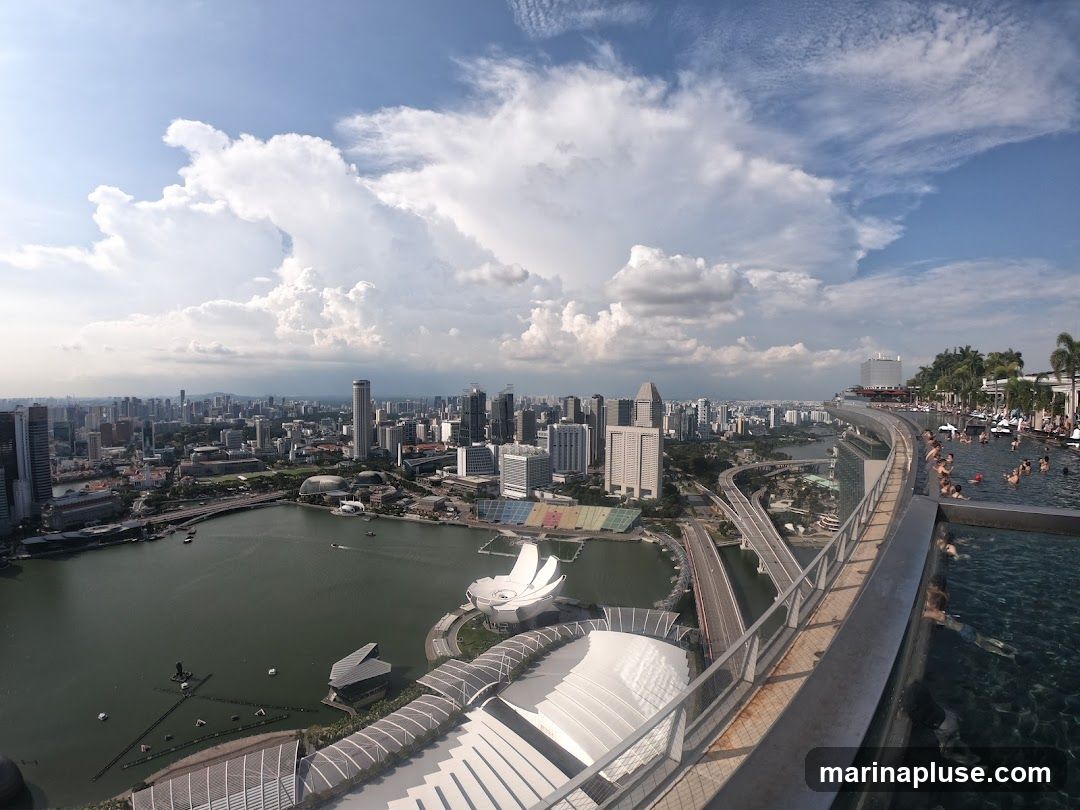 A wide-angle, panoramic view captures a bustling city skyline under a partly cloudy sky. In the foreground, an infinity pool with several people swimming and relaxing overlooks the cityscape. The pool area features lounge chairs and palm trees, suggesting a resort or hotel setting. Below the pool, a complex network of roads and bridges connects various parts of the city. In the middle ground, a large body of water, possibly a bay or river, is dotted with boats. Prominent architectural structures, including a distinctive building with a boat-like roof, are visible. The background is filled with numerous skyscrapers and urban development, stretching towards the horizon.
