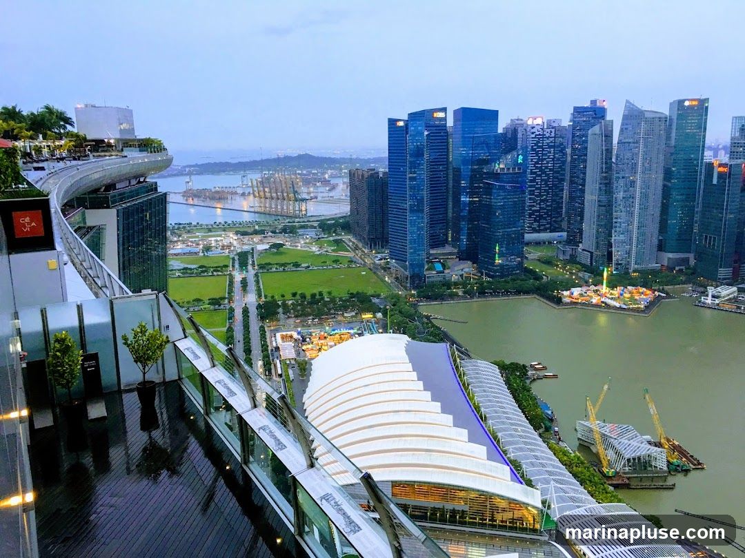 A panoramic view from a high vantage point, likely a rooftop bar or restaurant, overlooking the Singapore skyline. In the foreground, a sleek, modern outdoor seating area with glass railings and dark wooden decking is visible. Beyond this, the iconic Marina Bay Sands hotel and its integrated resort complex are prominent. The cityscape features numerous skyscrapers, a large body of water, and a distant view of a port with cranes. The sky is overcast, suggesting either early morning or late evening.