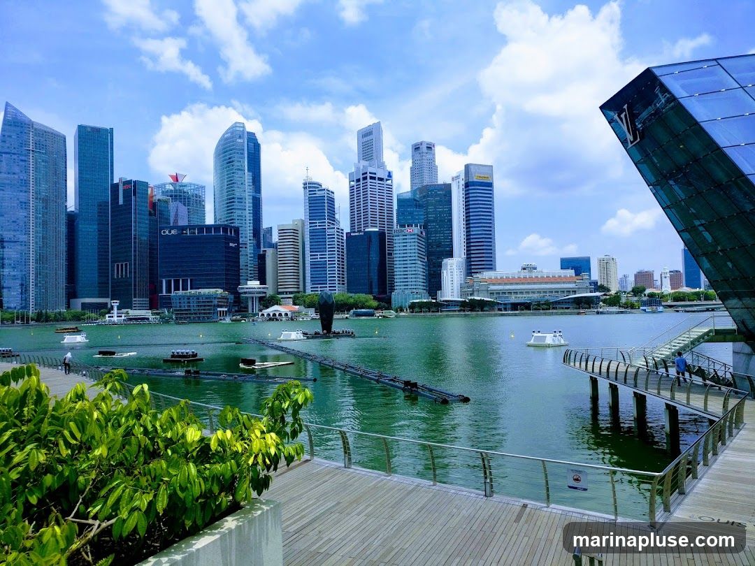 A panoramic view of the Marina Bay skyline in Singapore, featuring numerous modern skyscrapers and a body of water. In the foreground, a wooden boardwalk with lush green foliage leads towards the water. A distinctive glass-encased building with a prominent Louis Vuitton logo is visible on the right. The water is dotted with floating structures and a walkway extends over the water. The sky is blue with scattered white clouds.