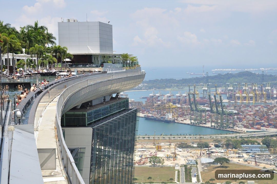 An aerial view of a rooftop infinity pool with people relaxing, overlooking a vast container port with numerous cranes and ships. The pool is situated on a modern building with lush greenery and lounge chairs. In the background, the expansive port stretches towards the horizon, with the ocean and distant islands visible under a partly cloudy sky.