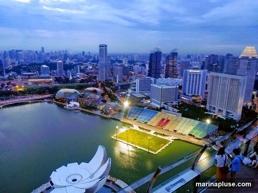 An aerial view of a city skyline at dusk, with a body of water in the foreground. Several modern skyscrapers dominate the background, while a stadium with colorful seating and a sports field is visible on the water. People are seen standing on a viewing platform in the lower right corner, looking out at the view. The sky is a deep blue with some clouds.