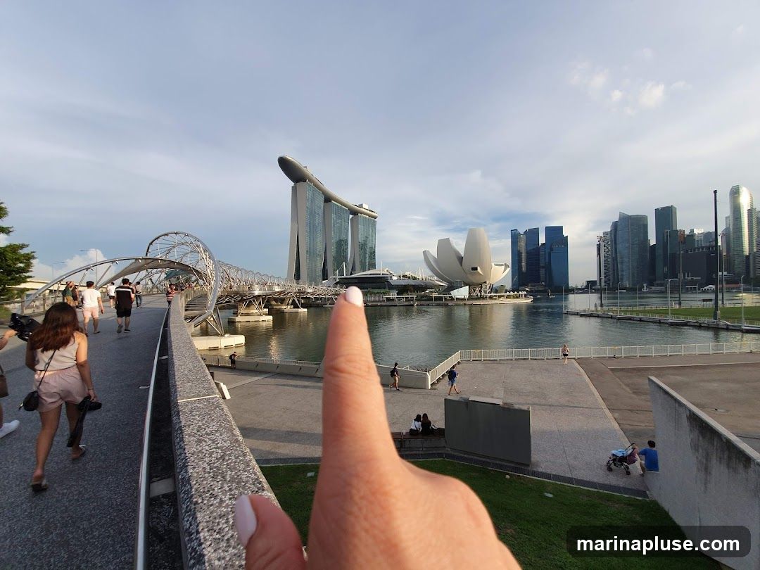 A wide-angle view of the Singapore skyline, featuring the iconic Marina Bay Sands hotel and the Helix Bridge. People are walking on the bridge and along the waterfront. The ArtScience Museum is also visible. The sky is partly cloudy, suggesting it might be late afternoon or early evening. A hand with a manicured fingernail is in the foreground, pointing towards the scene.