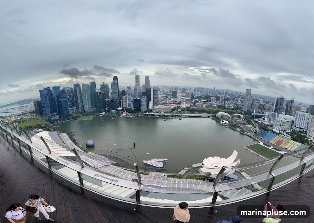 A wide-angle, fisheye lens view captures the Singapore skyline from the Marina Bay Sands SkyPark observation deck. The foreground features a wooden deck with several people observing the cityscape. In the mid-ground, the iconic Marina Bay Sands hotel structure is visible, along with the Supertree Grove and the ArtScience Museum. The background showcases a dense urban landscape with numerous skyscrapers and a body of water, likely Marina Bay. The sky is overcast with dramatic clouds.