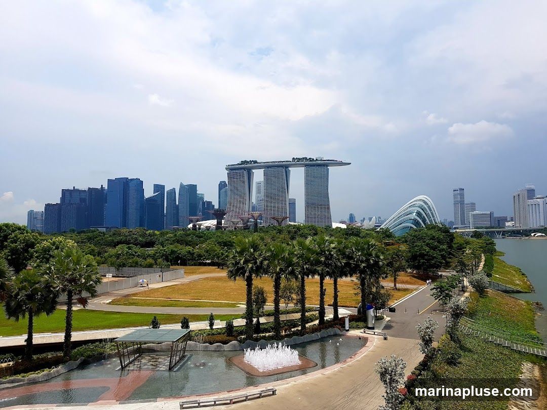 A panoramic view of the Singapore skyline, featuring the Marina Bay Sands hotel, the Supertree Grove, and the Flower Dome and Cloud Forest conservatories at Gardens by the Bay. In the foreground, a park area with palm trees, a water feature with fountains, and walking paths is visible. The Singapore River flows along the right side of the image. The sky is partly cloudy.