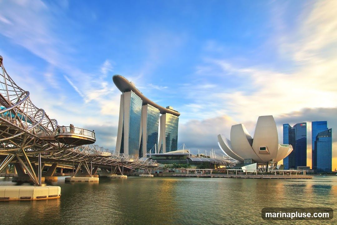 A wide shot of the Marina Bay Sands hotel and the ArtScience Museum in Singapore, with the Helix Bridge in the foreground. The sky is blue with wispy clouds, and the sun is setting, casting a warm glow on the buildings. The water in the bay reflects the sky and the architecture.