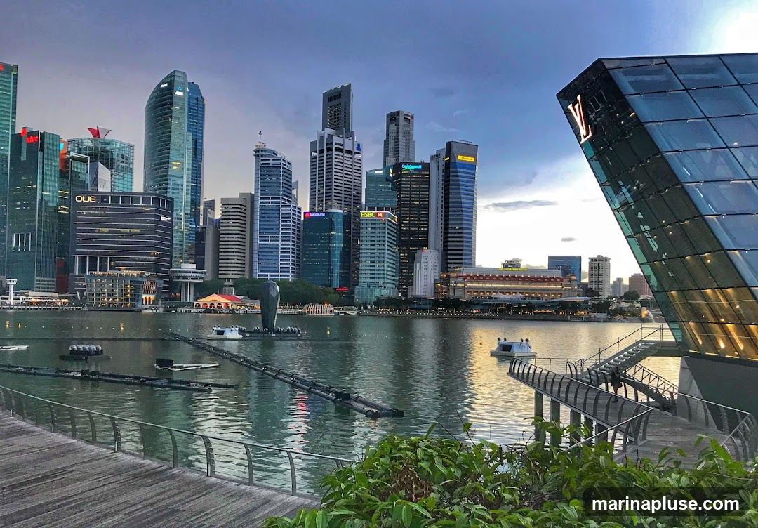 A wide shot of the Singapore skyline at dusk, with the Marina Bay Sands hotel and other skyscrapers in the background. A modern glass building with a Louis Vuitton logo is in the foreground on the right. The water in the bay reflects the city lights.