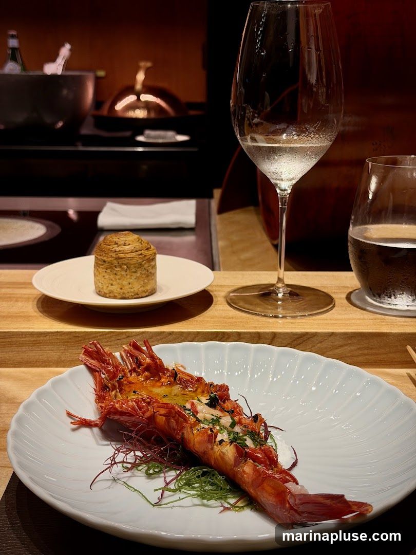 A close-up of a large, cooked langoustine or prawn served on a white, fluted plate. The seafood is garnished with herbs and sits on a bed of what appears to be white sauce and seaweed. In the background, a small pastry or bread roll is visible on a separate plate, along with a wine glass filled with water and another glass with a dark beverage. The setting suggests an upscale dining experience.