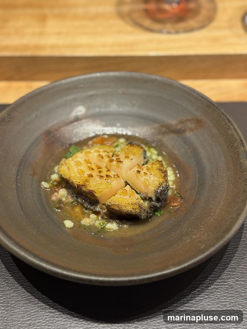 A close-up shot of a seared fish dish served in a rustic ceramic bowl. The fish, with its golden-brown crust and flaky interior, is presented on a bed of small, light-colored grains and finely diced vegetables, possibly tomatoes and herbs, in a light broth. The bowl has a textured, earthy glaze. In the blurred background, a wooden surface and a hint of a wine glass suggest a dining setting.