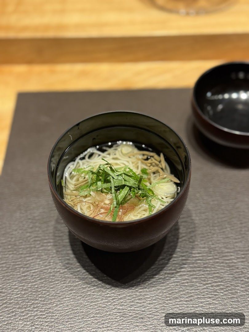 A close-up shot of a bowl of noodle soup, garnished with fresh green herbs and thinly sliced ginger. The soup is served in a dark, round bowl, placed on a textured placemat. Another empty dark bowl is visible in the background.