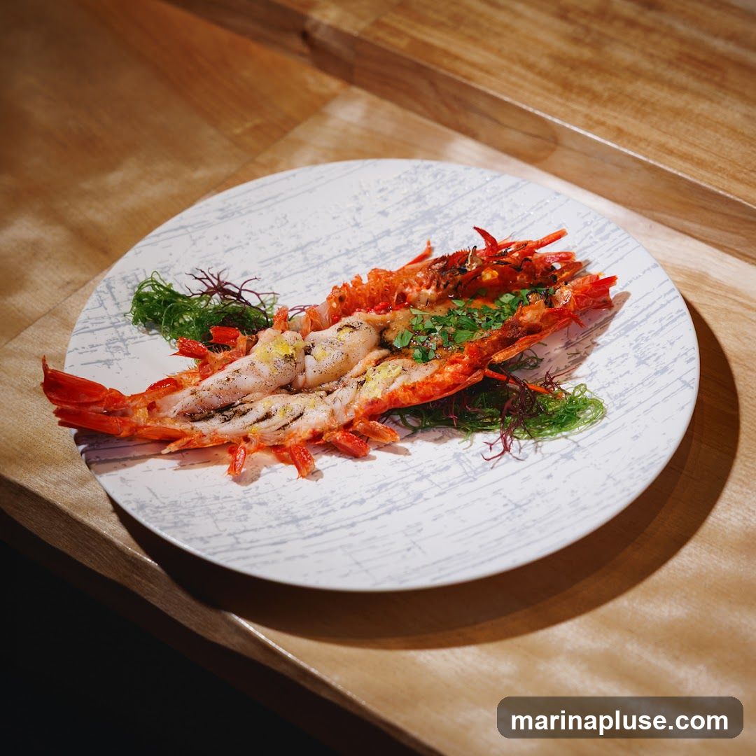 A whole lobster, split lengthwise and grilled, is served on a white plate with a grey abstract pattern. The lobster is garnished with chopped green herbs and served on a bed of seaweed. The plate is placed on a wooden surface.