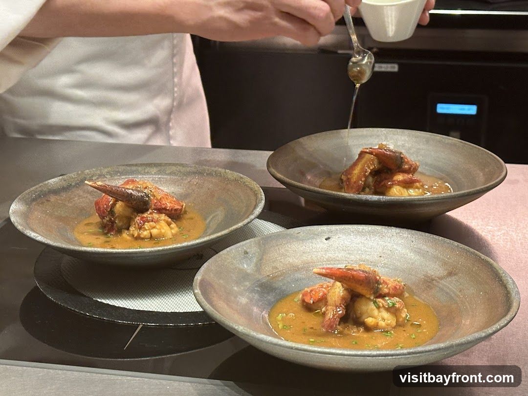 A chef in a white uniform is carefully drizzling a sauce over a beautifully plated lobster dish in a rustic bowl. The dish features succulent pieces of lobster in a rich, savory sauce, garnished with finely chopped herbs. Three bowls are visible, suggesting a multi-course meal or a shared appetizer. The background shows a professional kitchen setting.