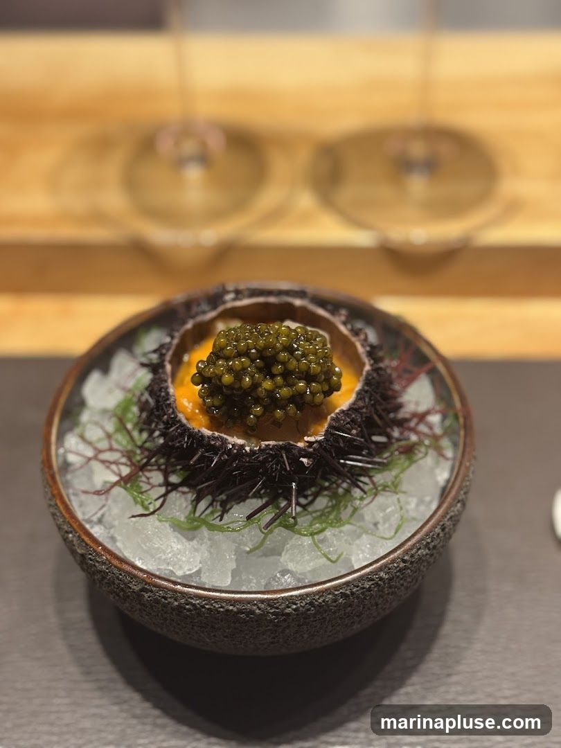 A close-up shot of a gourmet dish featuring sea urchin and caviar. The sea urchin shell is filled with bright orange uni and topped with a generous portion of dark caviar. It is presented on a bed of crushed ice and garnished with delicate seaweed. The dish is served in a textured, dark bowl, with blurred wine glasses visible in the background.