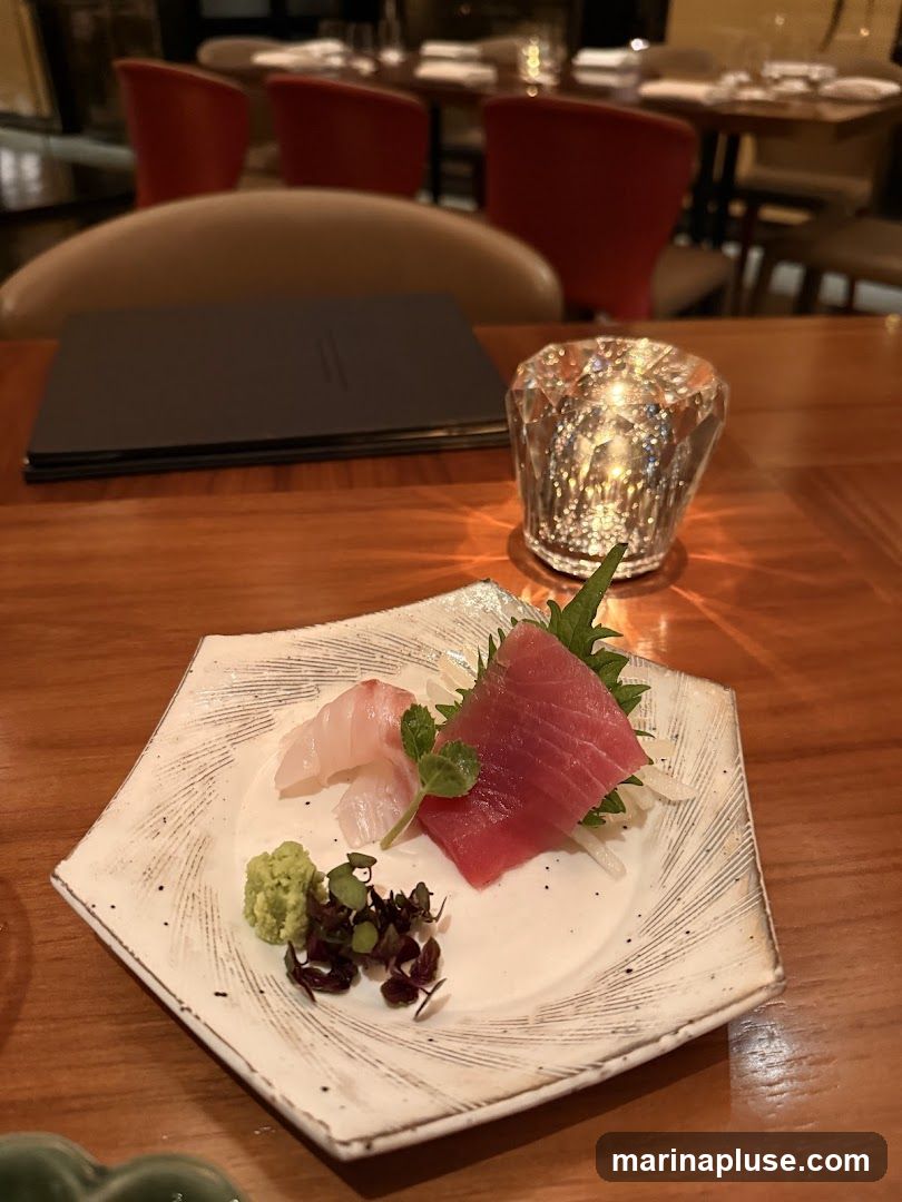 A close-up view of a sashimi platter featuring slices of red tuna and white fish, served with wasabi and microgreens on a textured white plate. A faceted glass candle holder with a lit candle sits to the right, casting a warm glow. In the background, a dark menu is visible on a wooden table, along with blurred red chairs and dining tables, suggesting a restaurant setting.