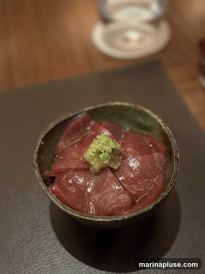 A close-up shot of a bowl filled with thinly sliced raw tuna sashimi, topped with a dollop of bright green wasabi. The dish is presented in a rustic, dark-rimmed bowl, resting on a dark placemat. In the blurred background, a white coaster and a hint of wooden table are visible.