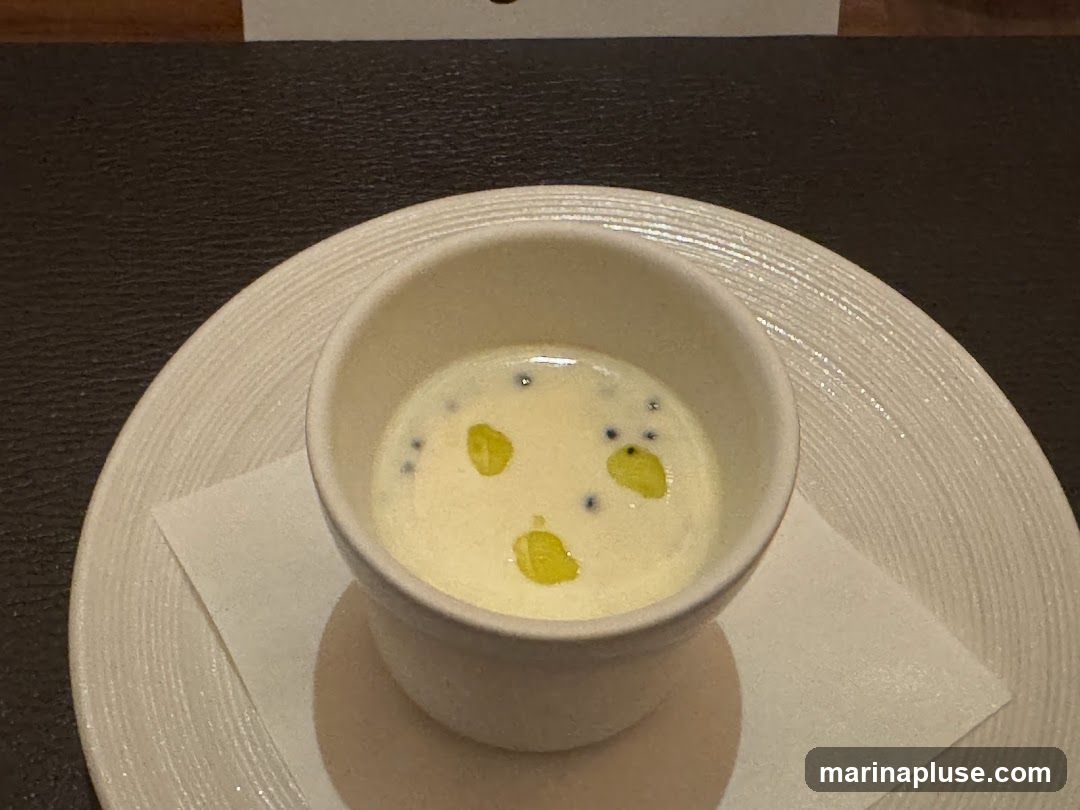 A small white bowl filled with a creamy white soup is placed on a textured white plate. The soup is garnished with three drops of yellow oil and several small dark blue or black beads. A white napkin is tucked under the bowl. The background is a dark textured surface.