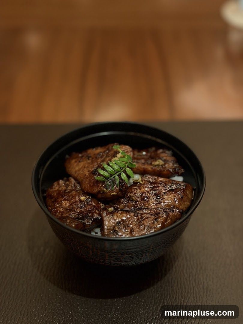 A close-up shot of a black bowl filled with rice and topped with several slices of grilled meat. The meat appears to be glazed and garnished with a sprig of green herbs. The background is blurred, showing a wooden texture.