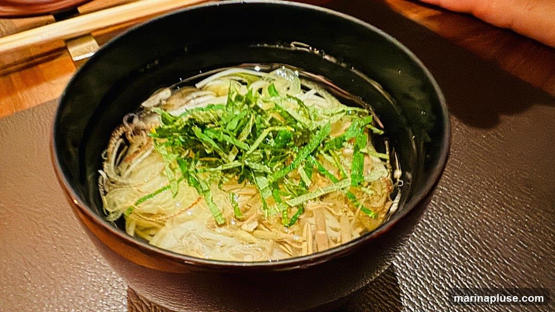 A close-up shot of a bowl of noodle soup, topped with finely chopped green herbs. The soup appears to be a clear broth with thin noodles and some other ingredients submerged. Chopsticks are visible in the upper left corner, and a person's hand casts a shadow on the right side of the image. The bowl is dark and sits on a textured brown surface.