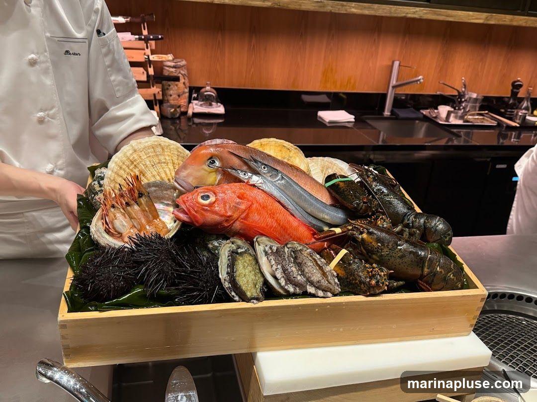 A chef in a white uniform presents a wooden tray filled with a variety of fresh seafood. The platter includes lobsters, sea urchins, abalone, scallops, shrimp, and several types of fish. The background shows a kitchen counter with a sink and wooden cabinetry.