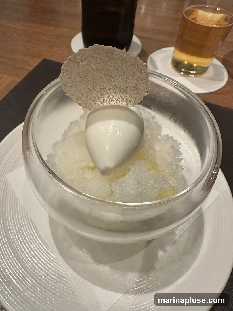 A close-up shot of a dessert served in a clear glass bowl. The dessert consists of a scoop of white ice cream or sorbet placed on top of a bed of crushed ice, possibly flavored with citrus or fruit. A thin, circular, textured garnish, resembling a crisp or wafer, is perched on the ice cream. The bowl sits on a white plate with a subtle ribbed pattern. In the background, two drinks are visible: one in a dark glass and another in a clear glass with a light-colored beverage, likely beer or cider. The setting appears to be a restaurant or cafe.