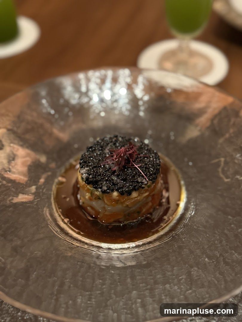 A close-up shot of a delicate seafood tartare dish, topped generously with black caviar and garnished with microgreens. The tartare is layered and served in a shallow, textured glass bowl with a rich, dark sauce. In the blurred background, a green beverage in a stemmed glass and a white coaster are visible, suggesting a restaurant setting.