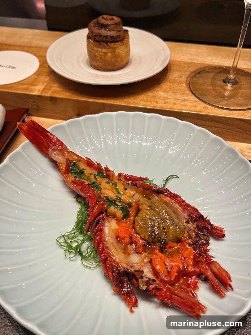 A close-up of a beautifully presented seafood dish, likely lobster or langoustine, served on a fluted pale blue plate. The seafood is split lengthwise, revealing succulent orange flesh and a rich, dark topping, possibly uni or a savory paste. It is garnished with finely chopped green herbs and served atop a bed of delicate green strands, resembling seaweed. In the background, a small, spiral-shaped pastry sits on a white plate, and a wine glass is visible. A coaster with faint text, possibly a restaurant name, is also present.