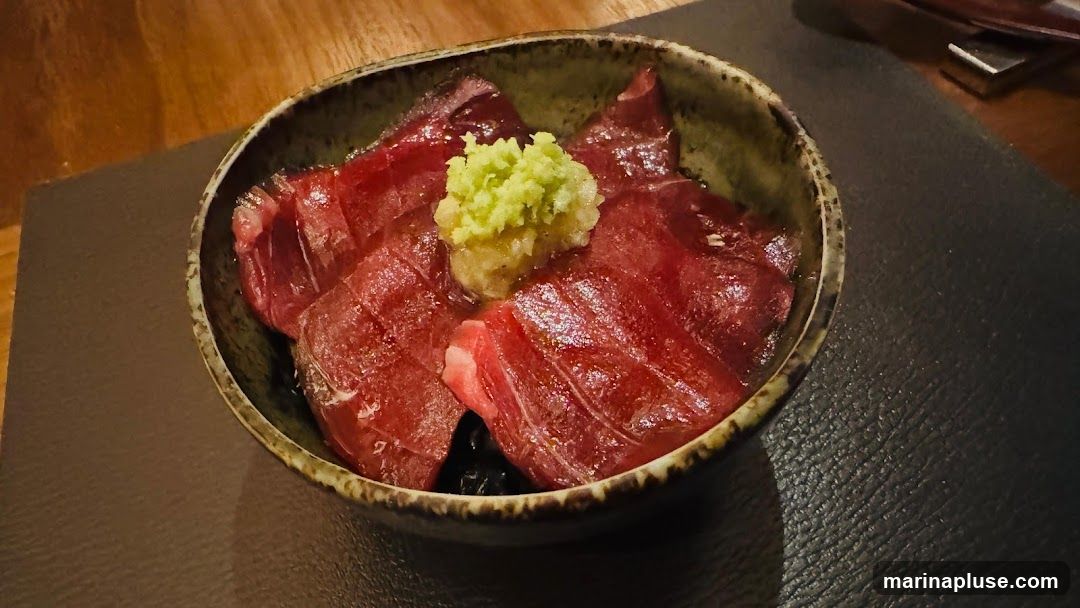 A bowl of tuna donburi, featuring thinly sliced raw tuna over rice, topped with a dollop of grated wasabi and ginger. The dish is served in a rustic ceramic bowl on a dark placemat.
