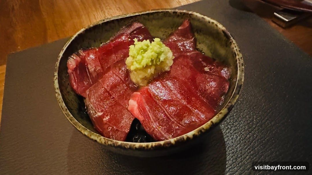 A bowl of tuna donburi, featuring thinly sliced raw tuna over rice, topped with a dollop of grated wasabi and ginger. The dish is served in a rustic ceramic bowl on a dark placemat.