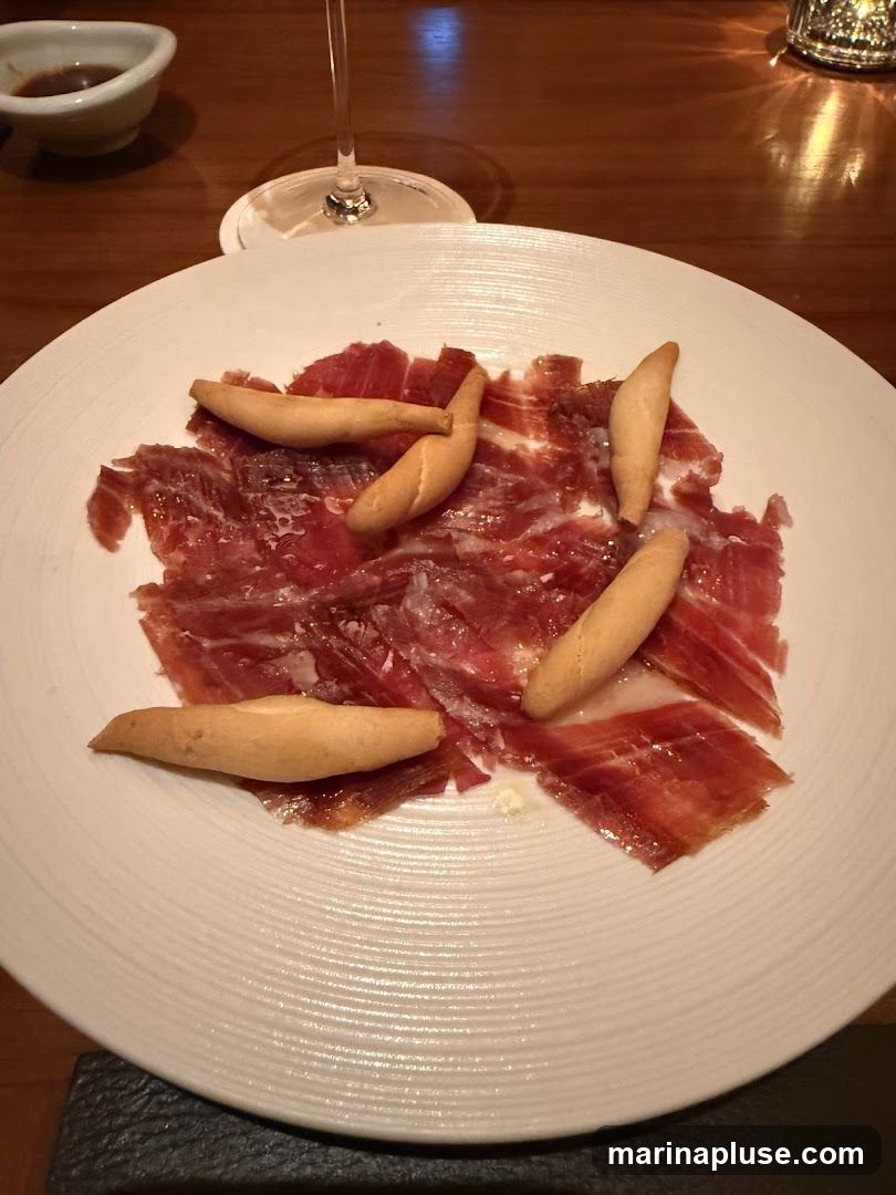 A white plate holds thinly sliced cured ham, garnished with several breadsticks. A small dish of dark sauce and a wine glass are visible in the background, set on a wooden table.