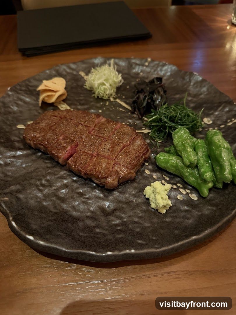 A close-up shot of a beautifully cooked steak, sliced and arranged on a dark, textured plate. The steak is accompanied by various garnishes, including what appears to be shredded daikon, pickled ginger, dark seaweed, green vegetables, and a dollop of wasabi. The meal is presented on a wooden table, with a stack of black menus visible in the background.