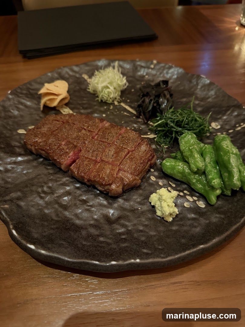 A close-up shot of a beautifully cooked steak, sliced and arranged on a dark, textured plate. The steak is accompanied by various garnishes, including what appears to be shredded daikon, pickled ginger, dark seaweed, green vegetables, and a dollop of wasabi. The meal is presented on a wooden table, with a stack of black menus visible in the background.