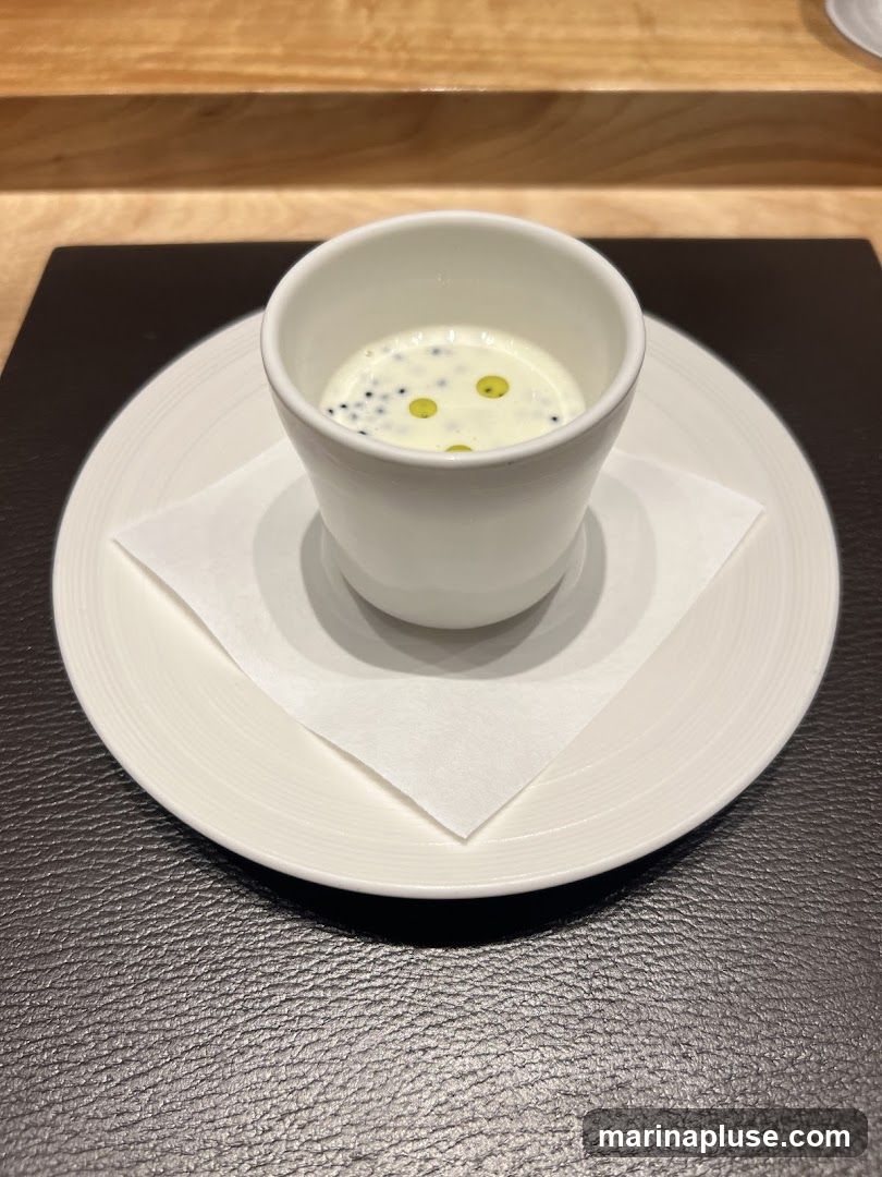 A close-up shot of a small white cup filled with a creamy white soup. The soup is garnished with black caviar and small droplets of green oil. The cup sits on a white saucer, which is placed on a dark textured placemat. In the background, a wooden surface is visible.