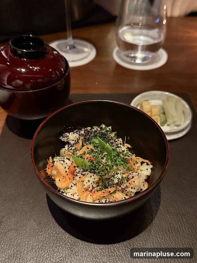 A close-up shot of a bowl of rice topped with small fried shrimp, black sesame seeds, and green herbs. A small dish of pickles is visible to the right, and a lidded dark red bowl is to the left. Two glasses, one with water and the other empty, are in the background.
