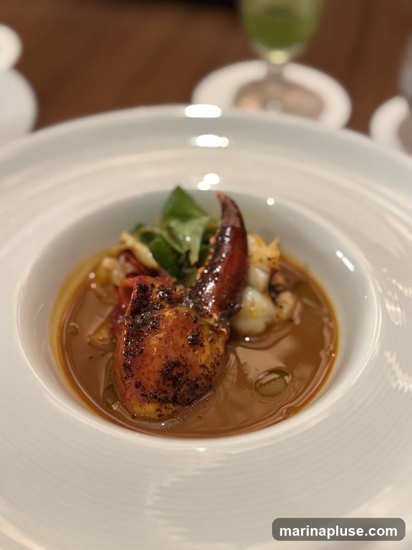 A close-up shot of a white bowl filled with a rich, brown broth. A single, cooked lobster claw with a dark red shell is prominently featured in the center, partially submerged in the soup. Small pieces of white seafood and green herbs are also visible within the broth. The bowl has a textured rim. In the blurred background, a green drink in a stemmed glass and other tableware can be seen.